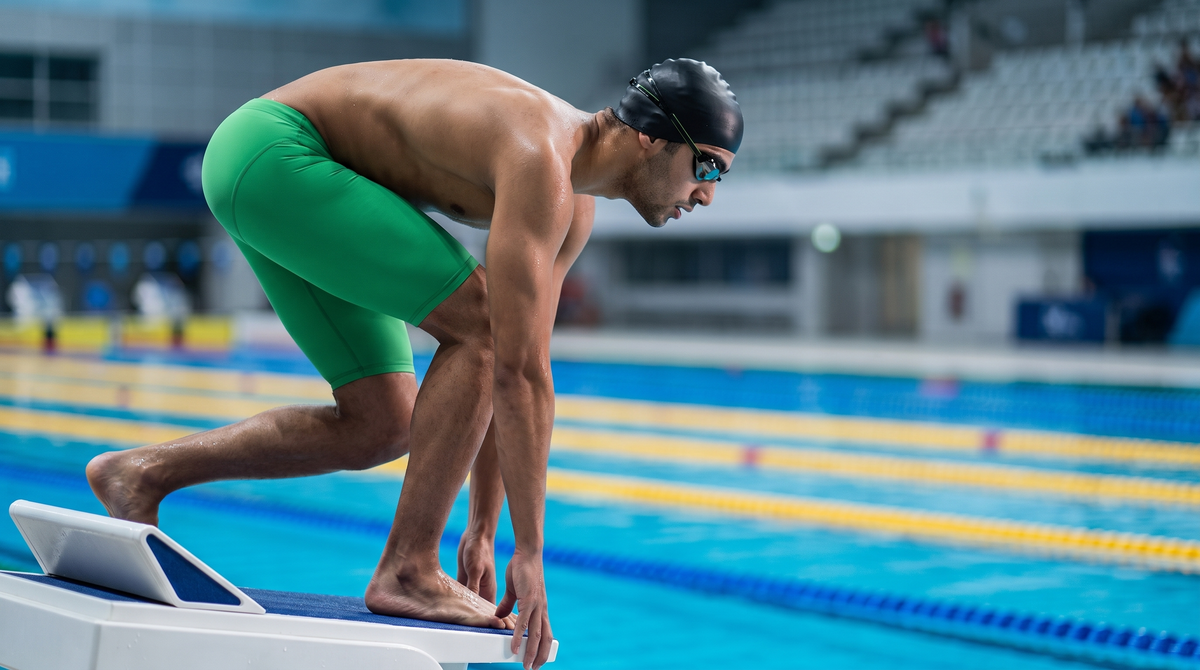 Swimmer in a green Mad Wave racing jammer and mirrored goggles on the starting block of a competition pool